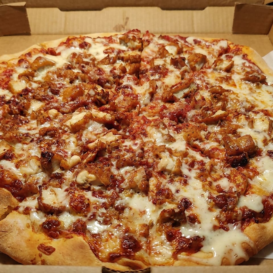 Large rectangular pizza topped with melted cheese, tomato sauce, sausage, and assorted roasted ingredients in a cardboard delivery box.
